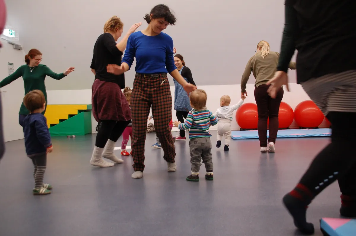 In einem Sportraum laufen mehrere Eltern mit Kleinkindern umher.