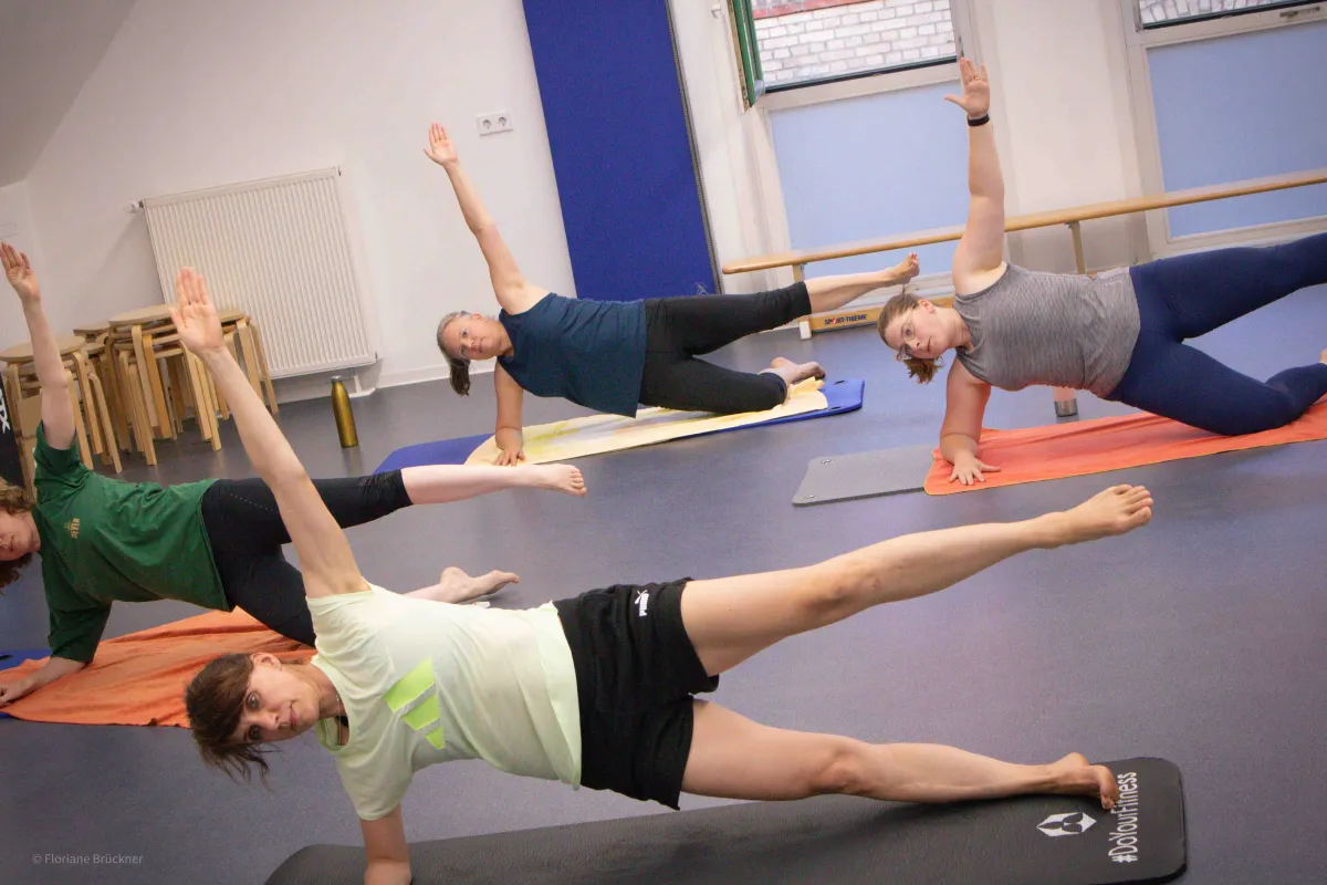 Mehrere Personen in einem Sportraum auf Gymnastikmatten machen eine Fitnessübung mit Arm in der Luft.
