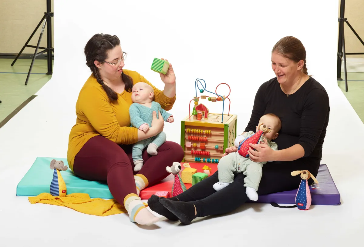 Zwei Frauen mit Baby auf dem Arm sitzen vor einer Foto-Leinwand, daneben stehen Spielgeräte.