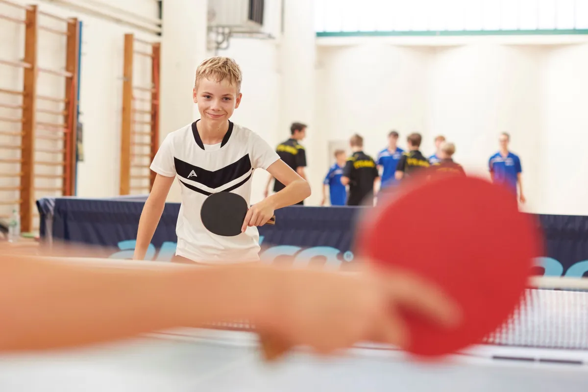 Im Vordergrund unscharf: Ein Arm hält eine Tischtenniskelle, im Hintergund scharf: Ein Junge mit Tischtenniskelle.