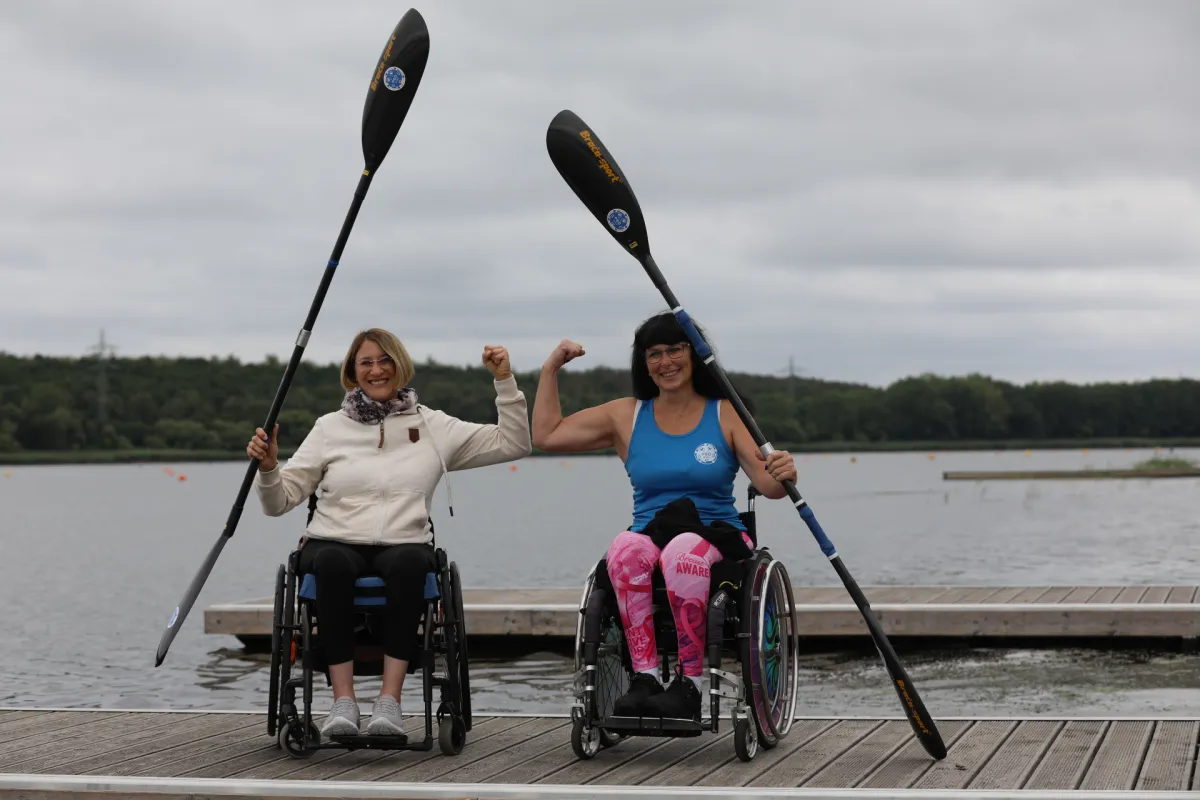 Zwei Personen im Rollstuhl mit Kajak-Paddel in der Hand auf dem Steg vor einem See.