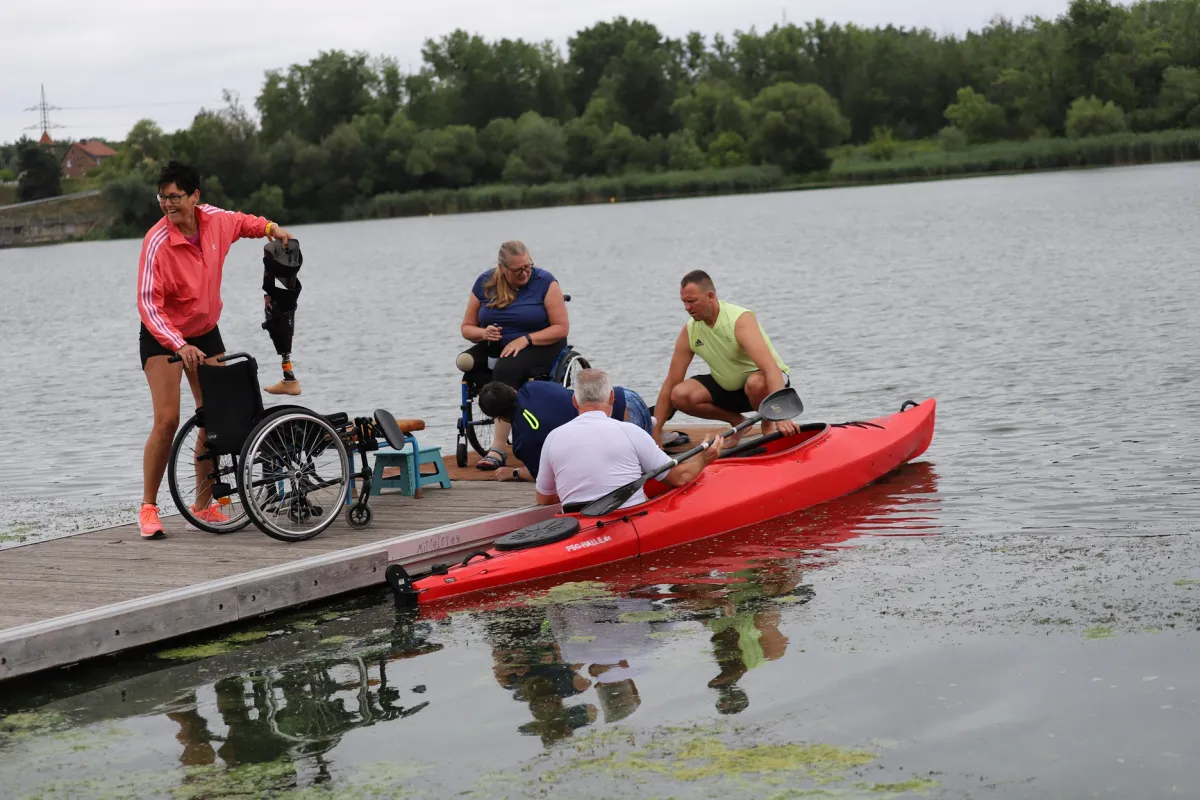 Para Kanu Wassercamp An einem Steg ist Trubel: Ein Mann sitzt im Kanu, Helfer an Land stehen neben einem Rolstuhl mit Beinprothesen und reden mit dem Mann.