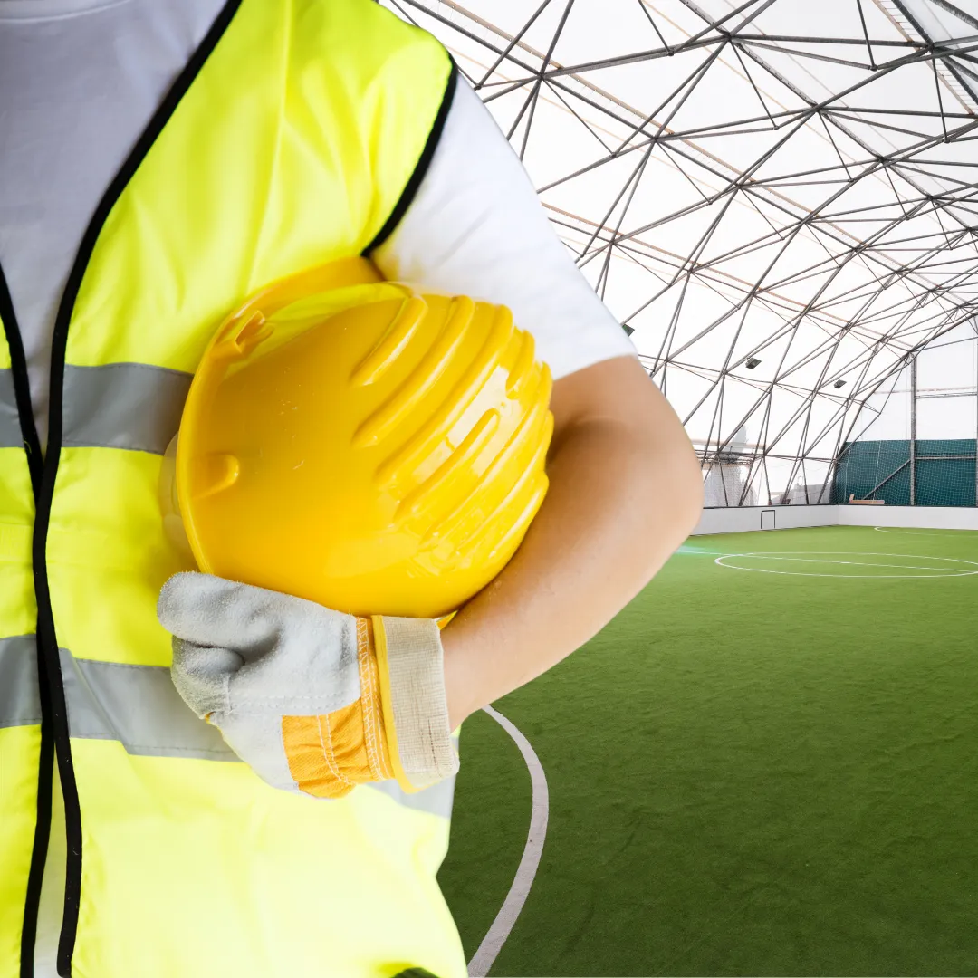 Sportstättenbau Im Hintergrund: Fußballhalle. Im Vordergrund: Oberkörper eines Bauarbeiters mit gelber Weste und gelbem Helm unterm Arm.