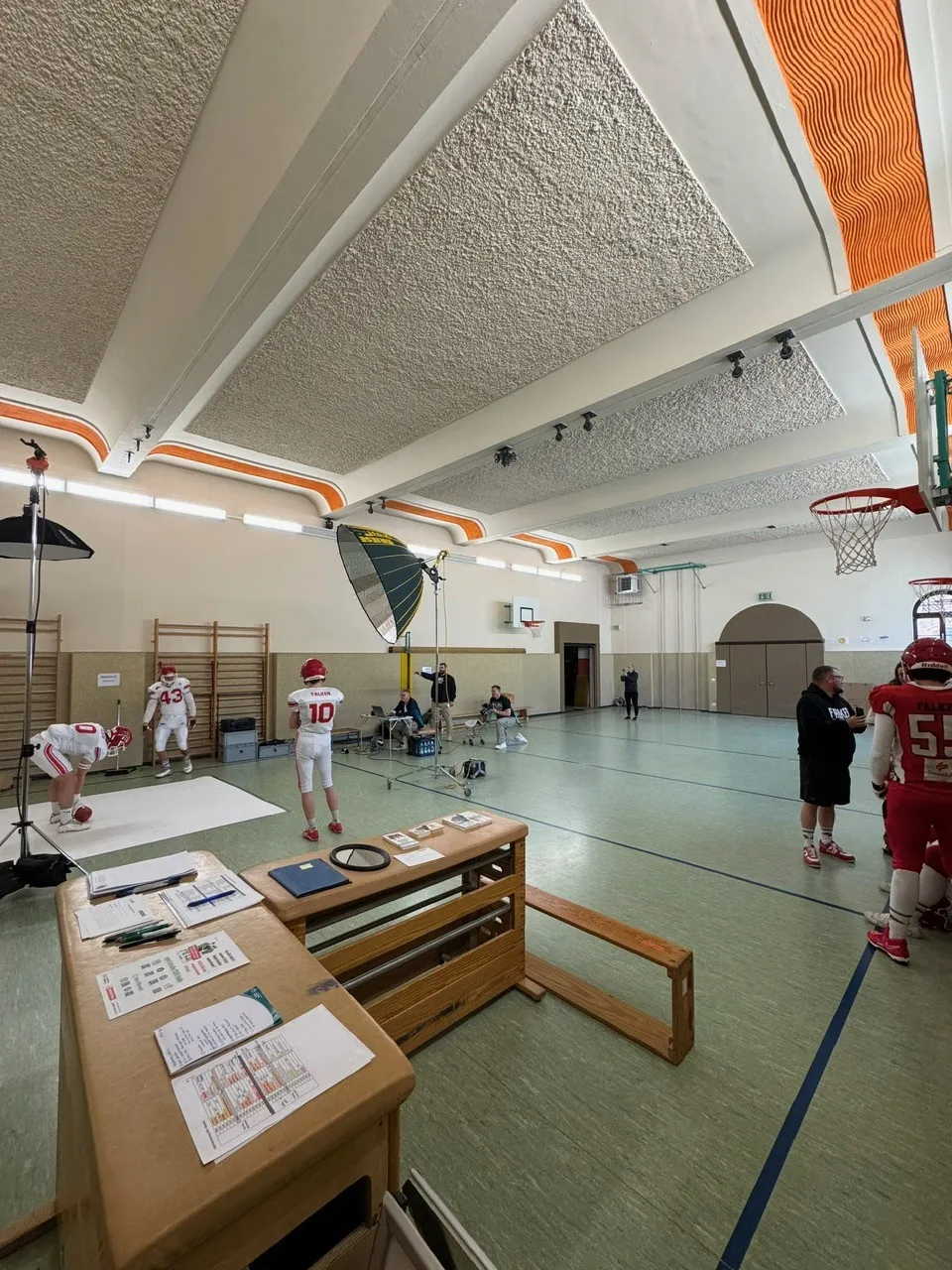 In einer Turnhalle ist ein Fotohintergrund aufgebaut, K&auml;sten mit Zetteln dienen als Anmeldung, Sportler laufen in der Halle umher,