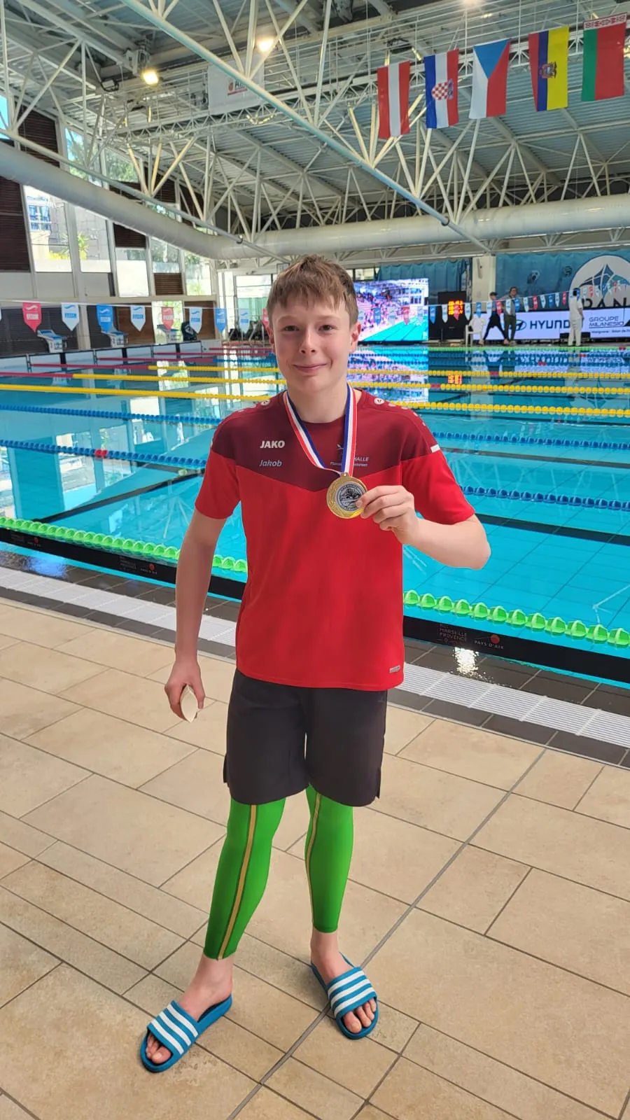 Junger Sportler zeigt stolz Medaille um den Hals vorm Schwimmbecken.