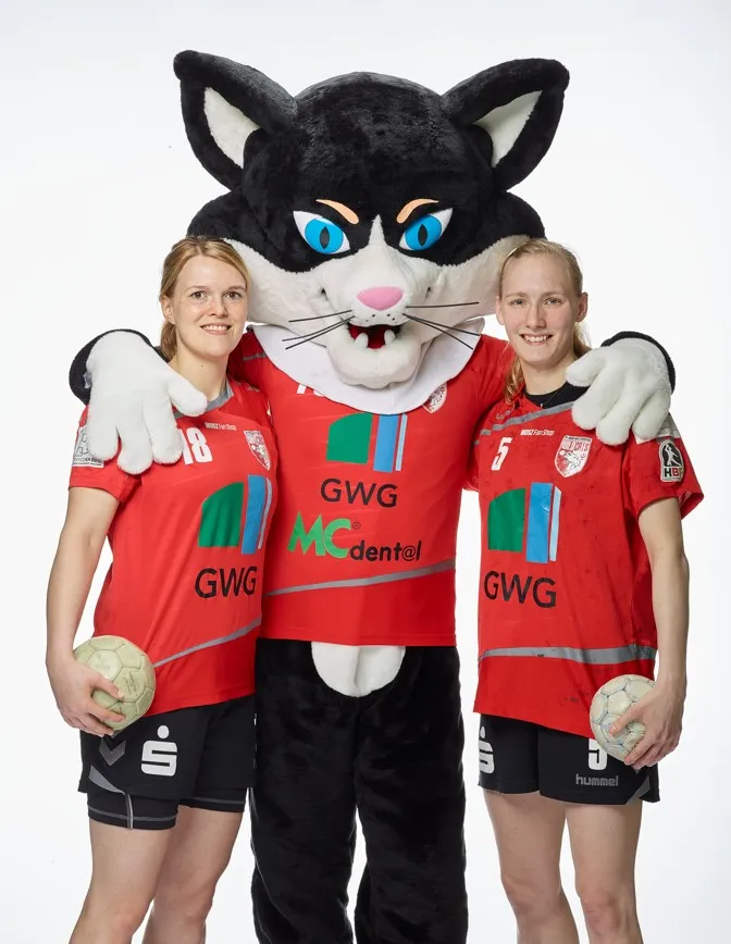 Wildcats-Maskottchen (eine Katze) mit jeweils einer Handballspielerin in beiden Armen.