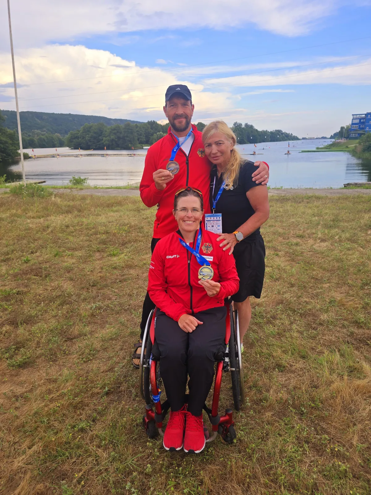 Frau im Rollstuhl und Mann jeweils in Trikots und mit Medaille, posieren mit weiterer Frau f&uuml;r Foto.