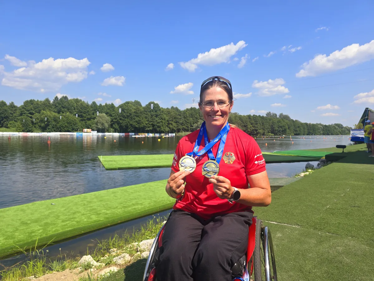 Frau im Rollstuhl in rotem Trikot mit Medaille um Hals vor Kanal