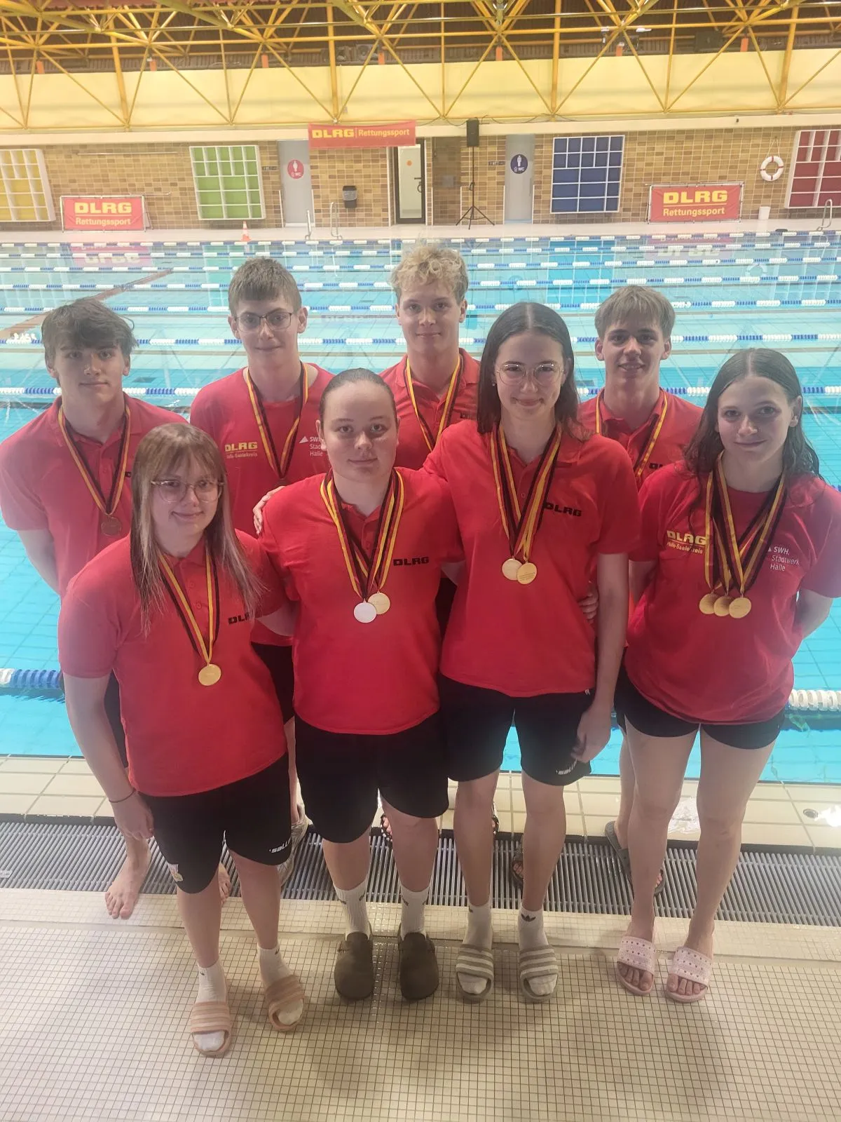 Acht Jungen und M&auml;dchen in roten Shirts mit Medaillen um den Hals vor Schwimmbecken.