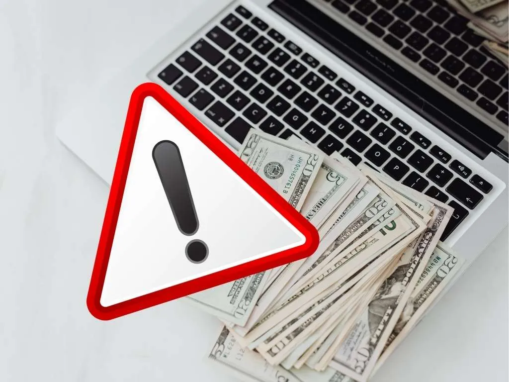 Geldscheine auf Laptoptastatur, davor Achtungschild mit rotem Rand und Ausrufezeichen