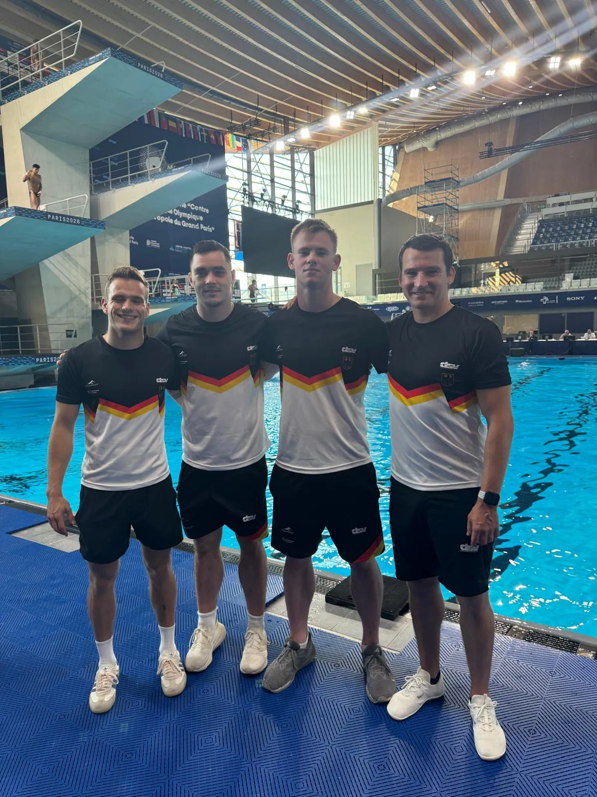Vier M&auml;nner in Deutschlandtrikots posieren vor Sprungturm in Schwimmhalle f&uuml;r Foto