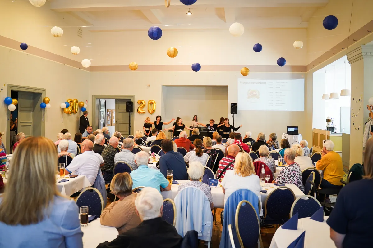 Menschen in mit Luftballons geschm&uuml;ckten Saal schauen auf Leinwand