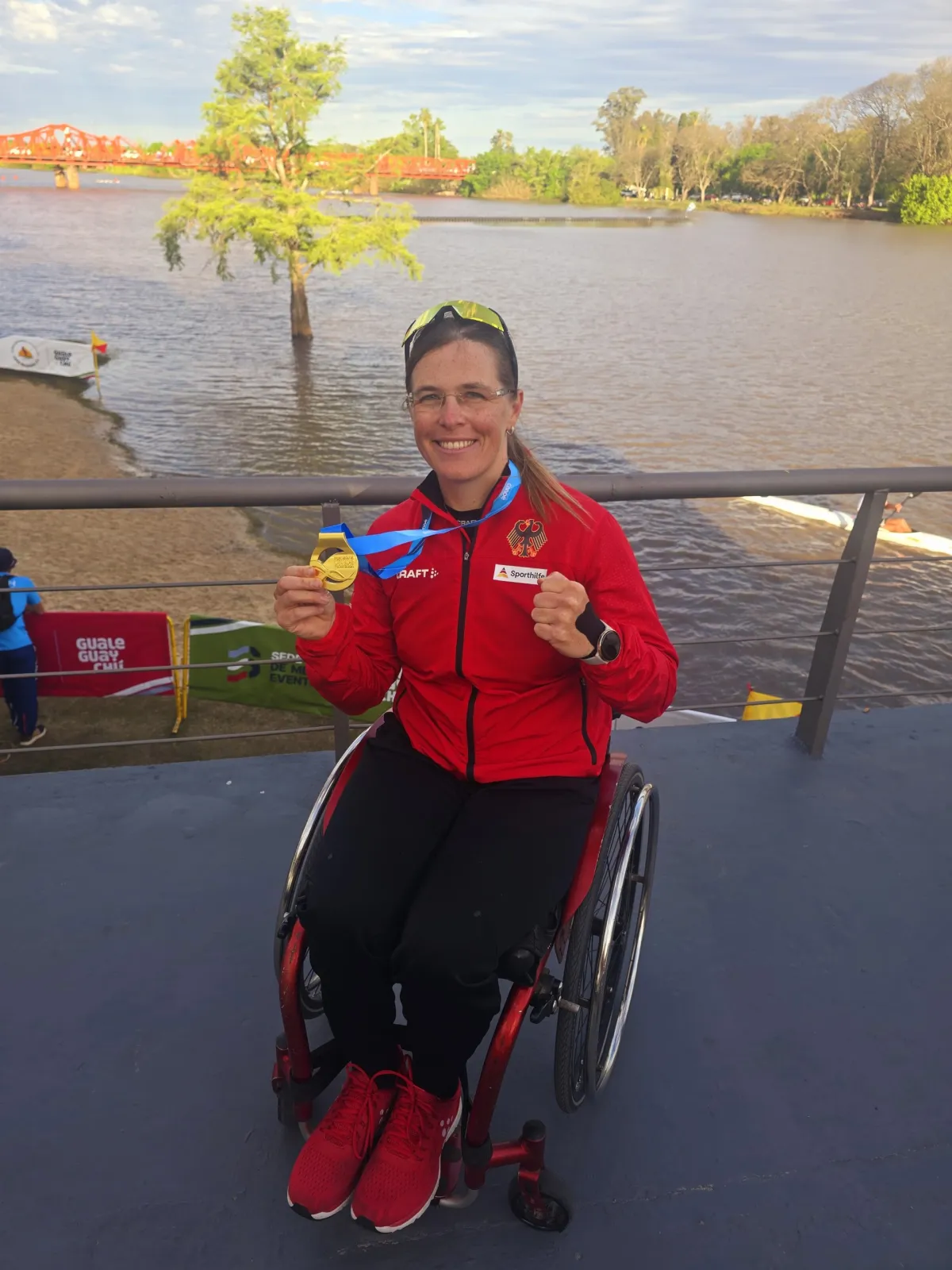 Frau in Rollstuhl in roter Sportjacke zeigt Medaille in Kamera