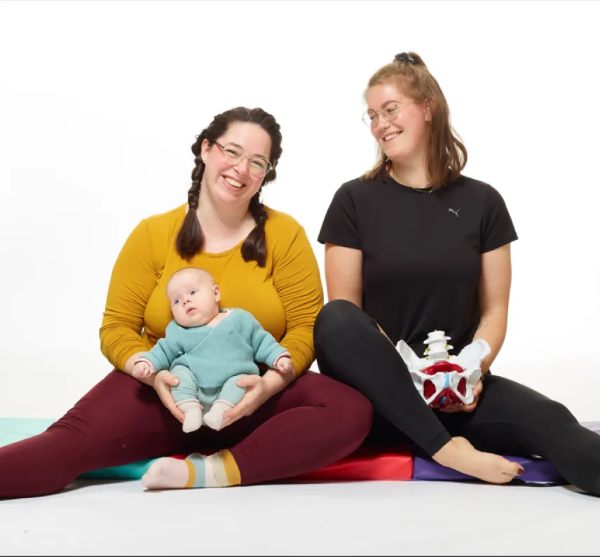 Zwei Frauen sitzen vor einer Fotoleinwand auf Gymnastikmatten, eine hat ein Baby auf dem Schoß.