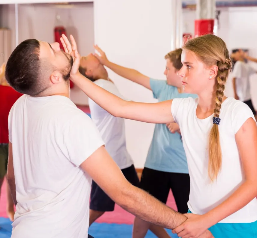Eine junge Dame mit Zopf hält einen jungen Mann am Handgelenk und gibt ihm im sportlichen Kontext einen Stoß gegen das Kinn.
