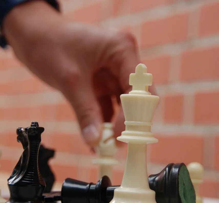 Ein weißer König steht auf einem Schachbrett, im Hintergrund schlägt eine Hand mit dem weißen Läufer die schwarze Dame.