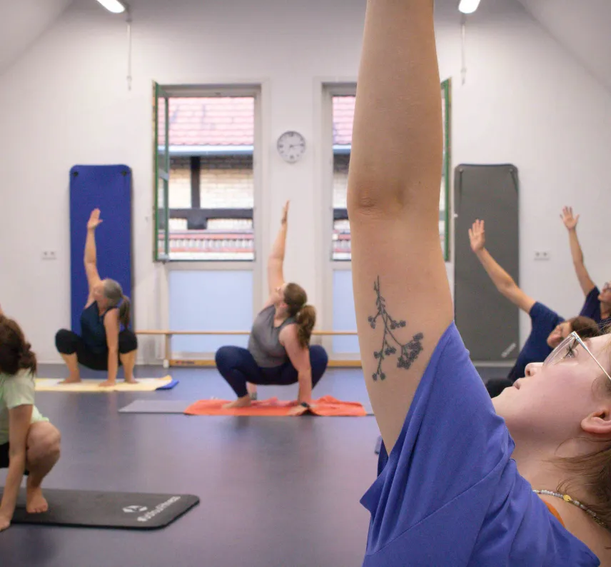 Mehrere Personen in einem Sportraum auf Gymnastikmatten machen eine Fitnessübung mit Arm in der Luft.