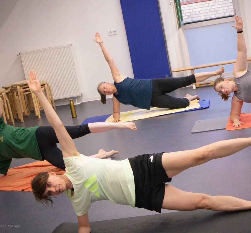 Mehrere Personen in einem Sportraum auf Gymnastikmatten machen eine Fitnessübung mit Arm in der Luft.