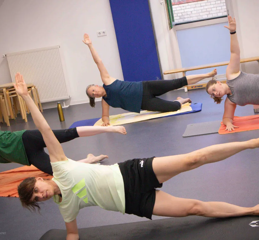 Mehrere Personen in einem Sportraum auf Gymnastikmatten machen eine Fitnessübung mit Arm in der Luft.