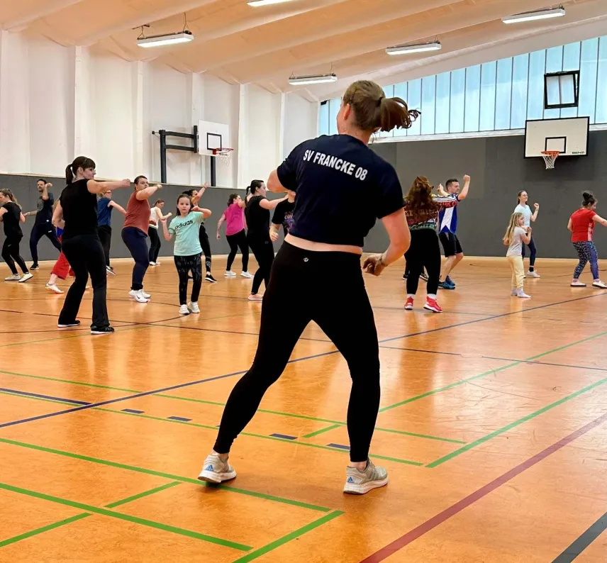 Vordergrund: Eine Frau in Vereinskleidung macht eine Tanzpose. Im Hintergrund sind viele Personen in einer Turnhalle, die sie nachmachen.
