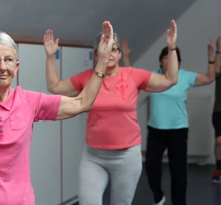 Mehrere Personen in einem Sportraum machen eine Gymnastikübung im Stehen.