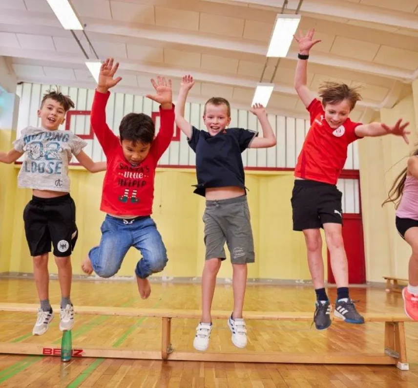 Fünf Kinder hüpfen fröhlich in einer Turnhalle von einer Bank.