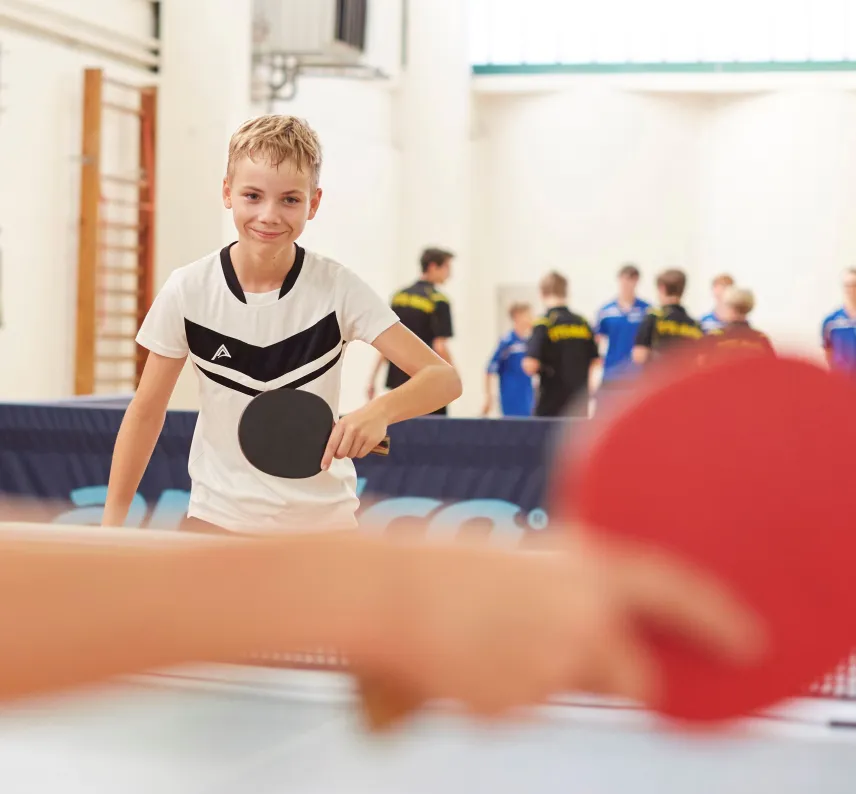 Im Vordergrund unscharf: Ein Arm hält eine Tischtenniskelle, im Hintergund scharf: Ein Junge mit Tischtenniskelle.