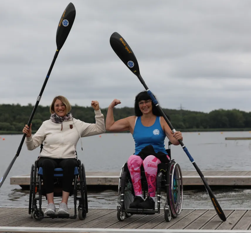 Zwei Personen im Rollstuhl mit Kajak-Paddel in der Hand auf dem Steg vor einem See.