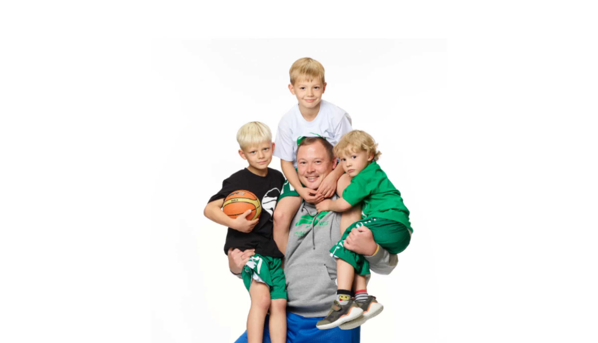 Ein Mann im Hoodie hat drei Jungen im Trikot auf seinen Schultern, den rechten Fuß auf einem Basketball.