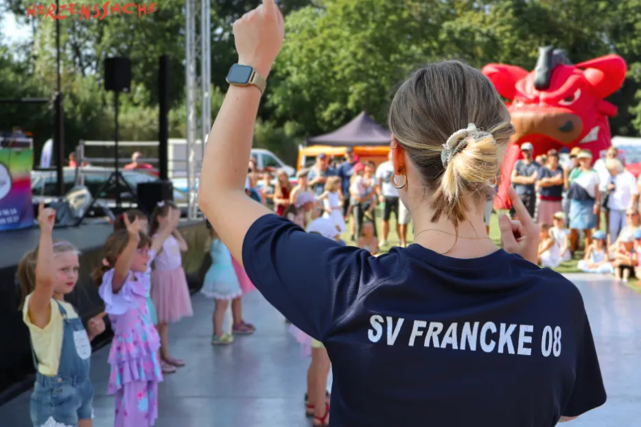Im Vordergund: Rückansicht einer Frau im SV Francke-Shirt. Sie hebt den Zeigefinger. Hintergrund: Kinder zanzen auf einer Tanzfläche in Formation.