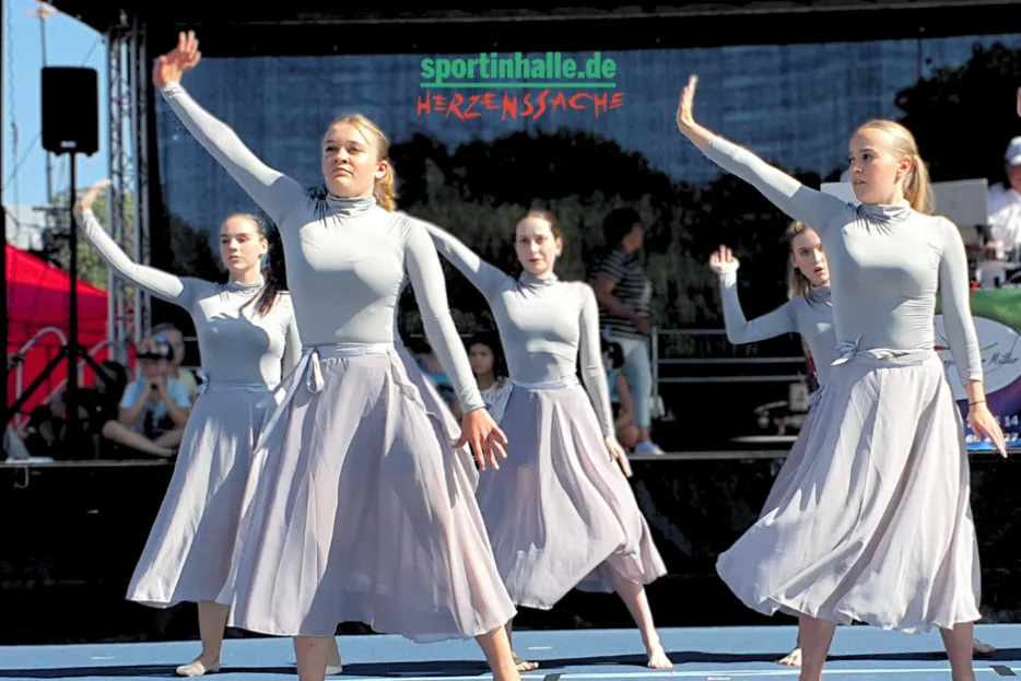 Fünf Frauen in fliederfarbenen Kleidern stehen in Tanzformation mit dem linken Arm in der Luft.