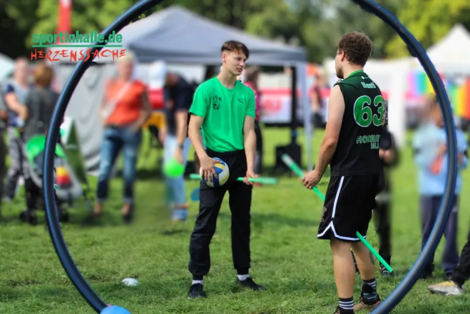 Fotogtrafiert duch einen Ring vom Quidditsch/Quadball sieht man zwei Spieler in grünen Trikots auf Besenstilen, einer hält einen Ball.