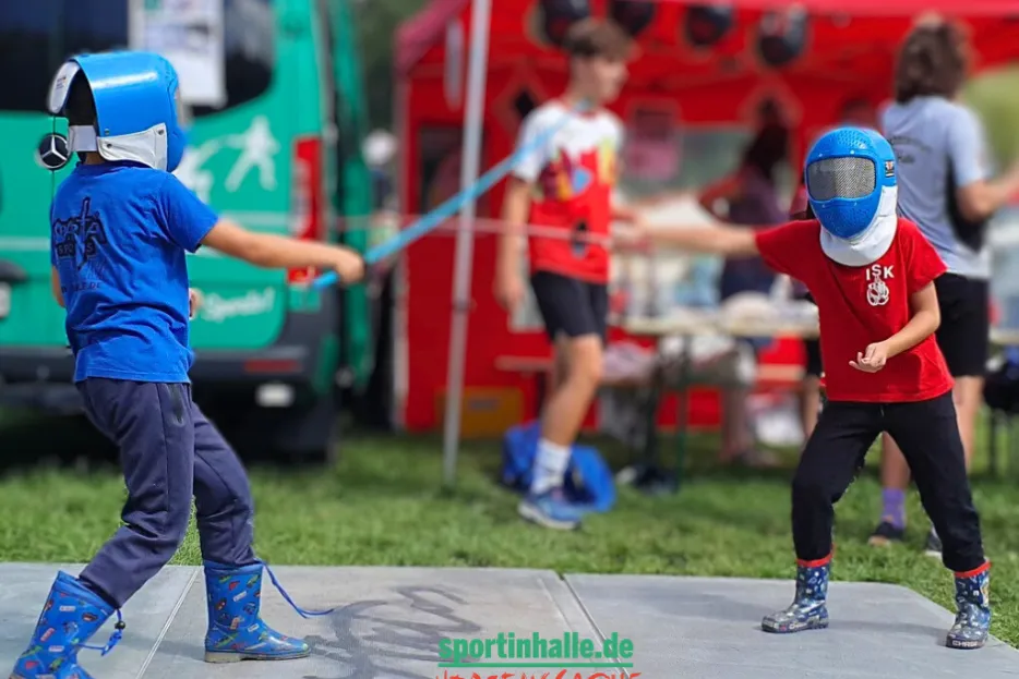 Zwei Kinder in Fecht-Masken mit Sportwaffen fechten auf einer Matte.