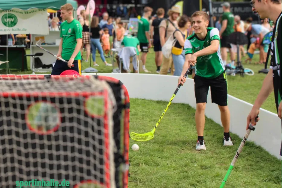 Unscharf im Vordergrund: Ein Floorballtor. Scharf im Hintegrund: Ein Spieler zielt mit Schläger und Ball aufs Tor.