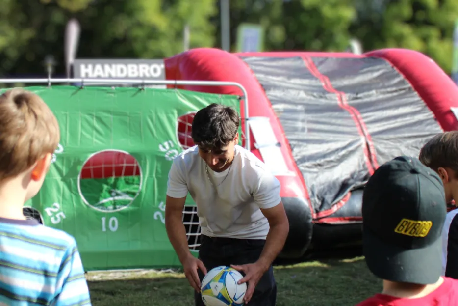 Ein junger Mann legt einen Fußball vor einer Torwand zurecht. Drei Jungen beobachten ihn dabei.
