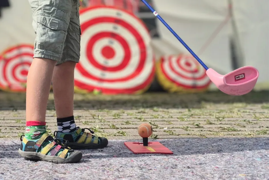 Bildausschnitt eines Jungen, der mit Golfschläger vorm Ball ausholt, um ihn auf eine Zielscheibe zu schießen.
