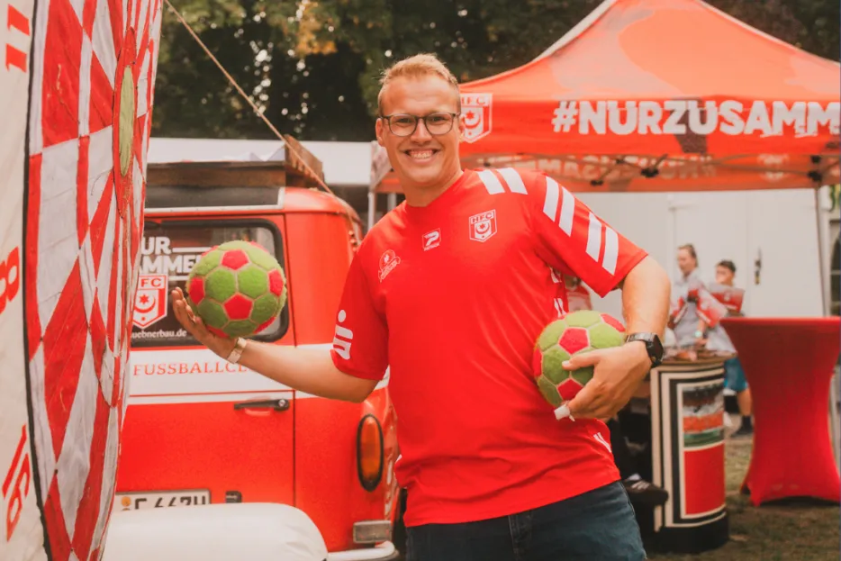 Ein Mann im roten T-Shirt hält einen Ball im Arm, einen anderen wirft er Richtung Kamera.