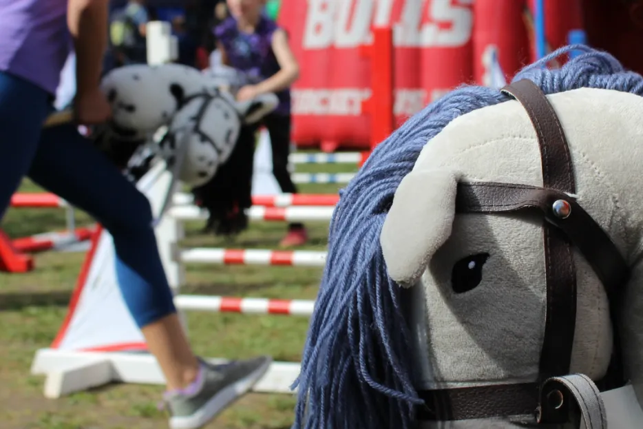 Im Vordergrund sieht man den Kopf einen Steckenpferdes, im Hintergrund springen Mädchen mit Steckenpferden über Sprünge.