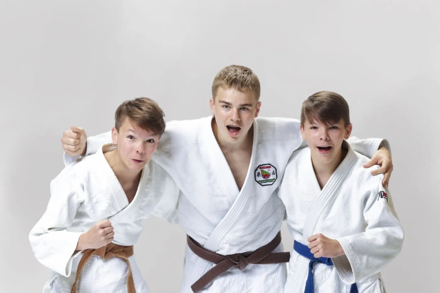 Drei junge Judoka mit Judogi-Jacke posieren Arm in Arm für die Kamera.