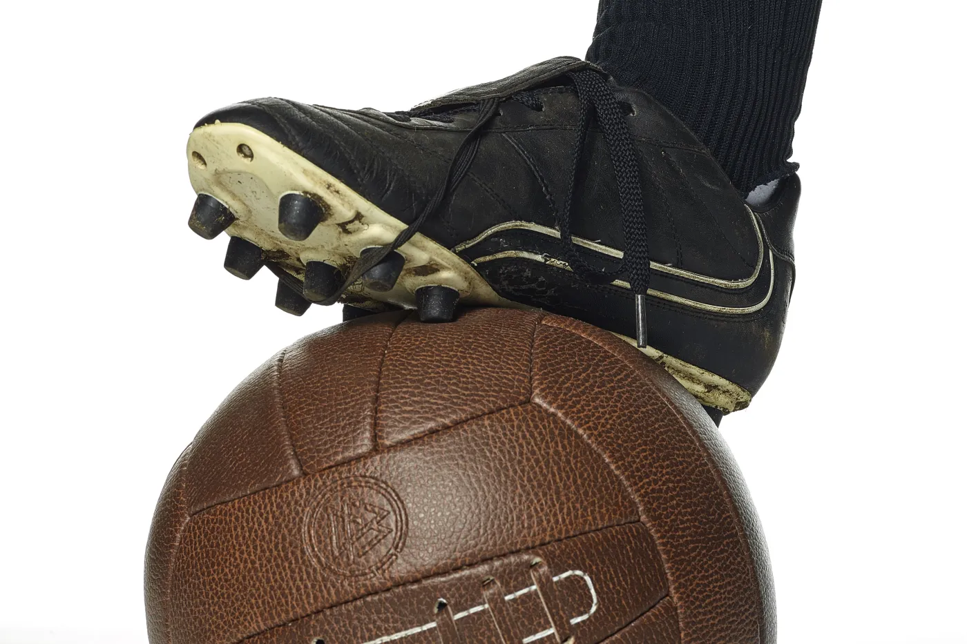 Zu sehen ist ein Bildausschnitt mit einem Fuß mit Fußballschuh, der einen alten Leder-Fußball annimmt.