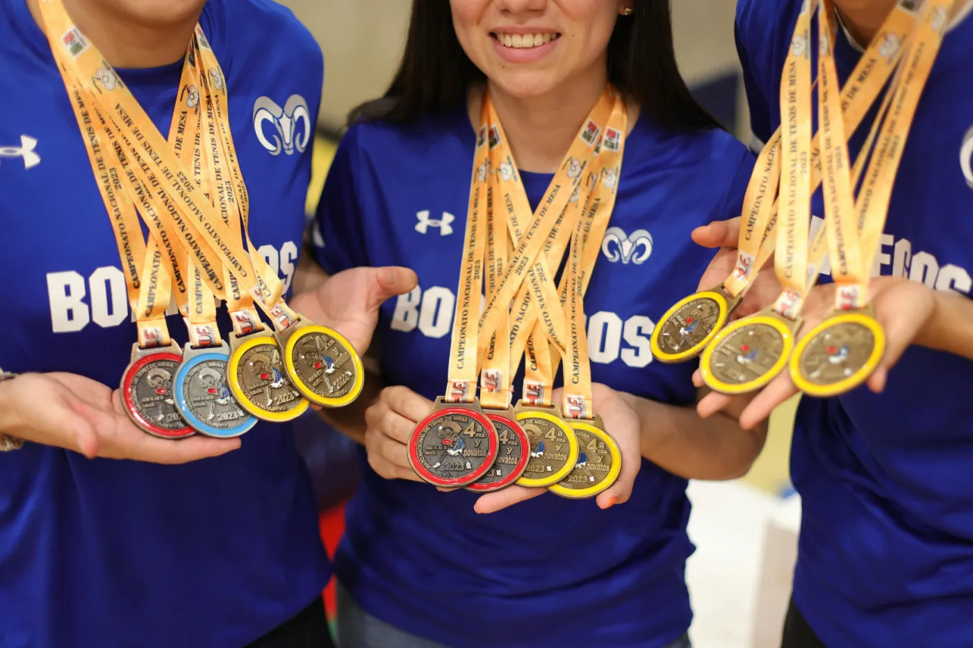 Drei Sportlerinnen in blauem Trikot halten jeweils 4 Medaillen um den Hals ins Bild.