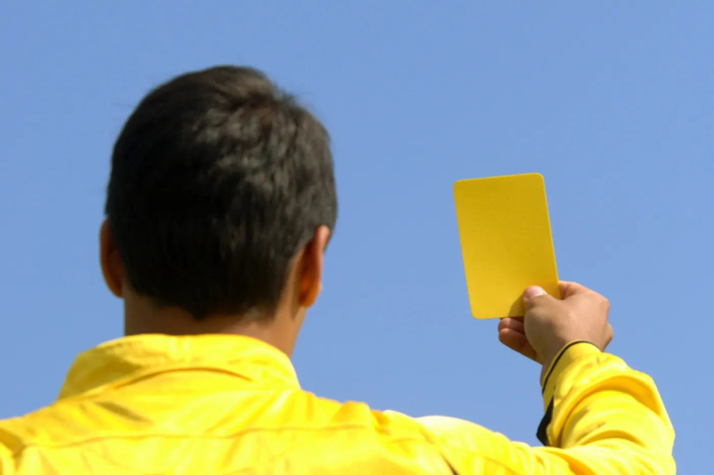 Zu sehen ist ein schiedsrichter in gelbem Trikot von hinten, wie er eine gelbe Karte zeigt.
