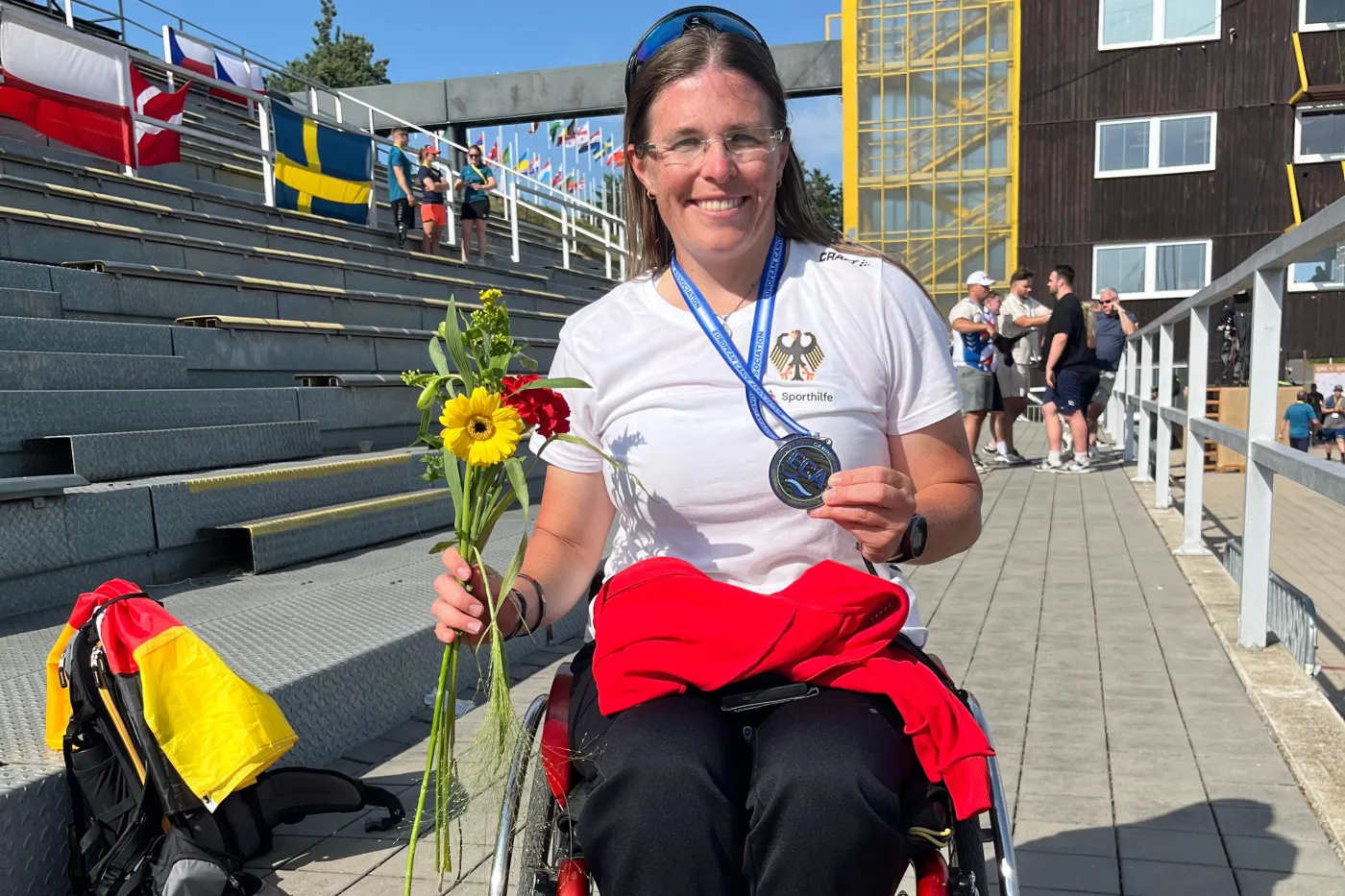 Anja Adler mit Bronzemedaille um den Hals und Blume in der rechten Hand im Rollstuhl auf dem Gelände der EM-Regattastrecke.