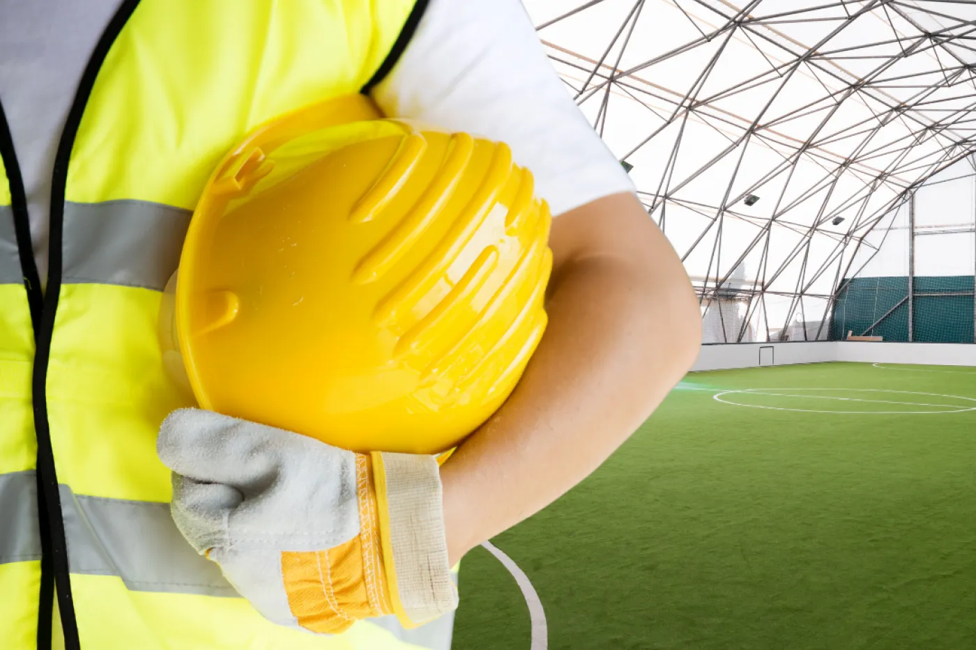 Symbolbild Sportstättenbau Zu sehen ist der Oberköper eines Bauarbeiters mit Warnweste und gelbem Hekm im Arm, im Hintegrrund ist eine Fußballhalle.