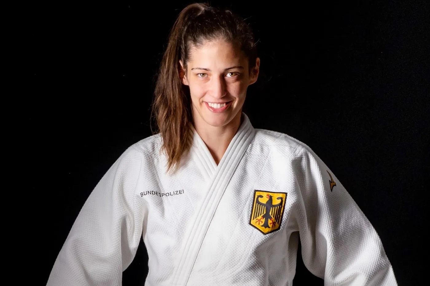Miriam Butkereit. Miriam Burkereit steht im weißen Judo-Anzug vor dunklem Hintergrund, den Bundesadler auf der Brust. Sie zieht ihren gürtel zurecht.