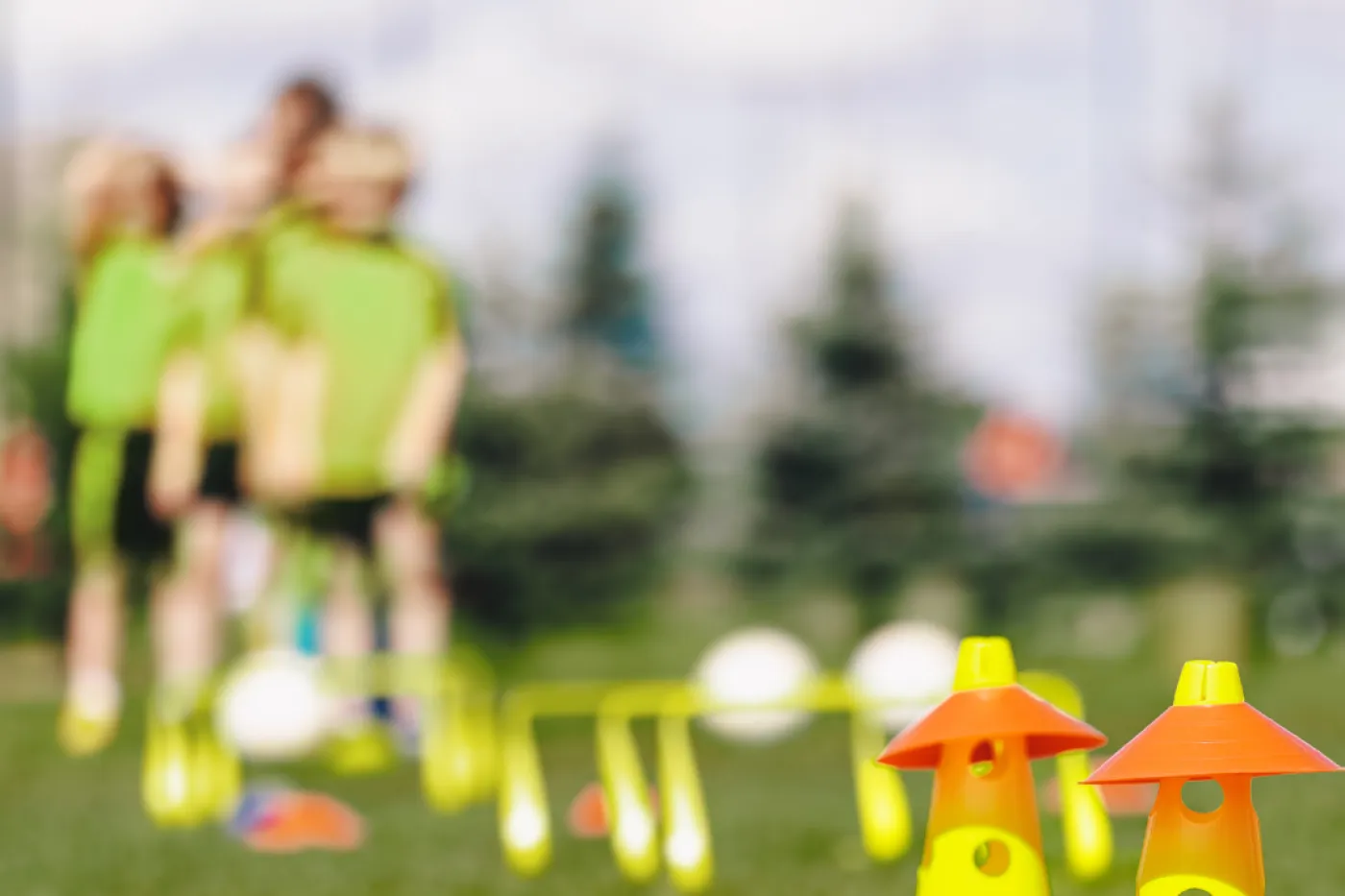 Kindersport im Vordergrund scharf: Gelbe Kegel und Reifen auf einer Wiese, im Hintergrund unscharf Kinder mit Bällen und grünen Trikots.
