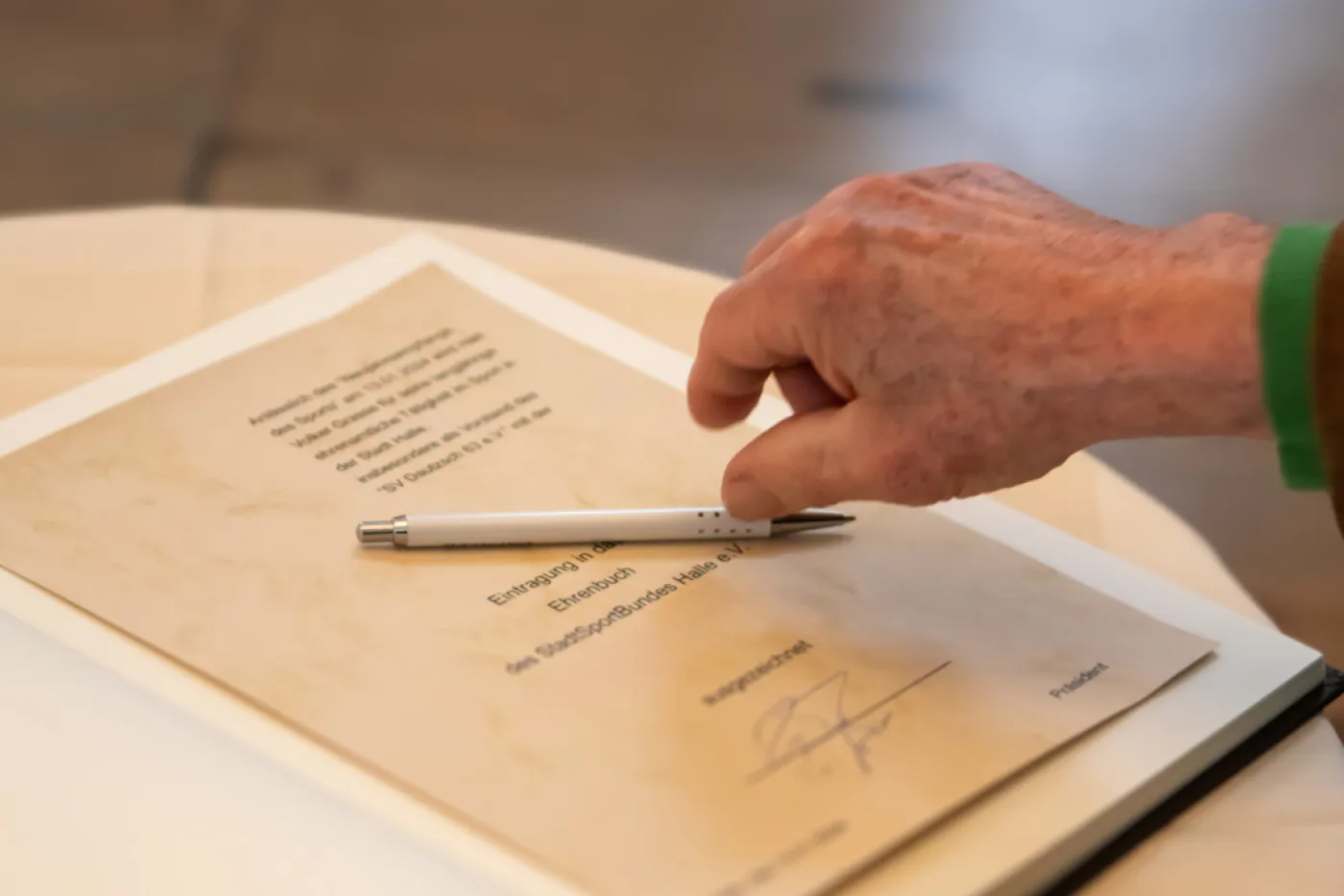 Ein Buch ist aufgeschlagen, eine Urkunde klebt darin. Eine Hand greift nach dem Stift auf der Urkunde.