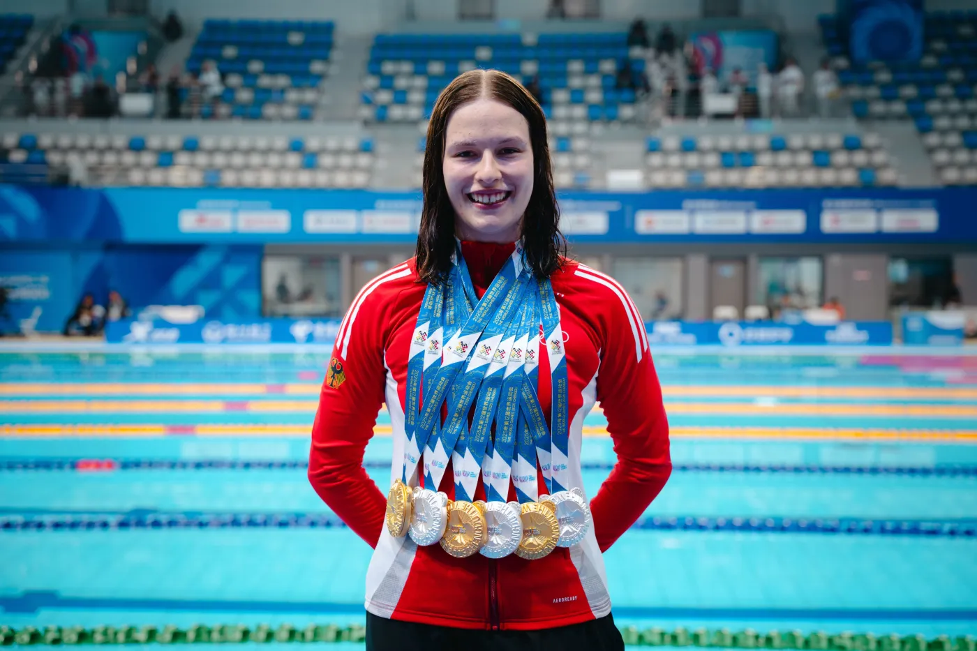 Lena Oppermann in roter Trainingsjacke mit drei goldenen und drei silbernen Medaillen um den Hals in der Schwimmhalle.