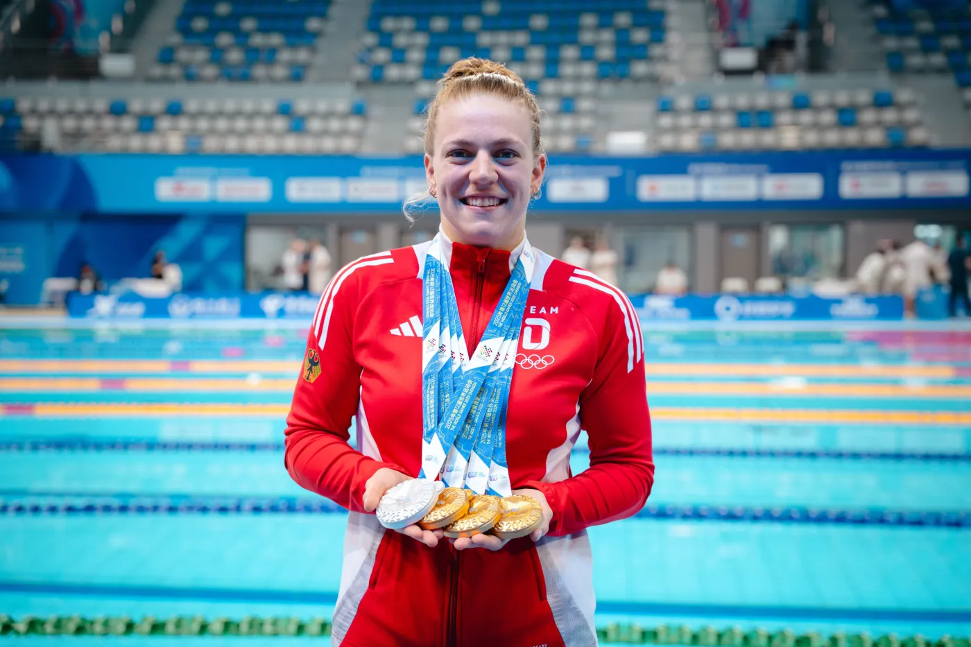 Undine Lauerwald Undine Lauerwald in roter Trainingsjacke mit drei goldenen und einer silbernen Medaille um den Hals in der Schwimmhalle.