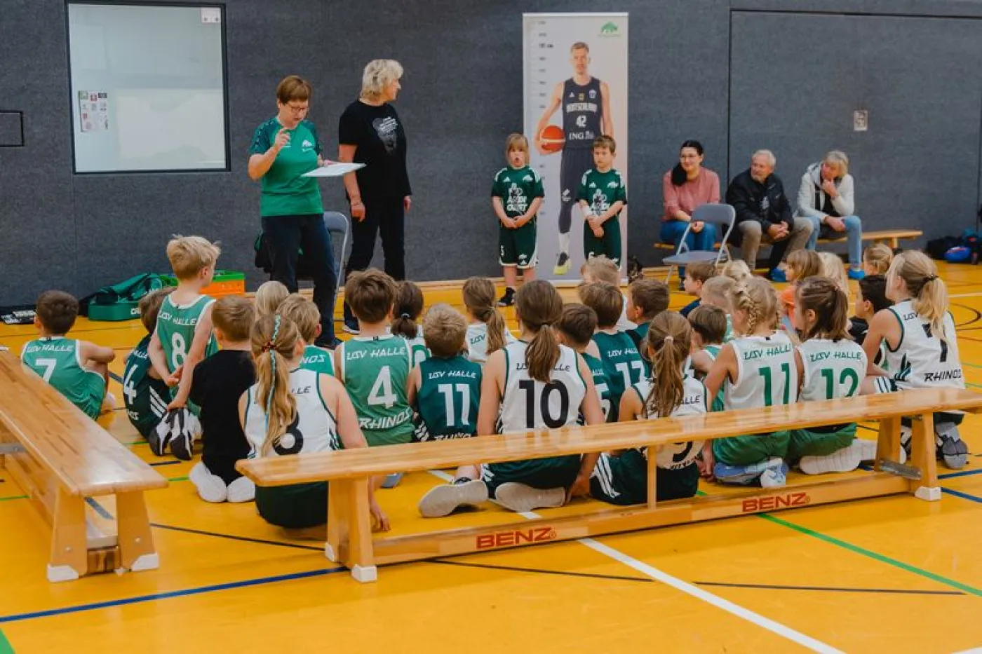 Kinder sitzen auf einer Bank und beobachten zwei von ihnen, die etwas erklärt bekommen.